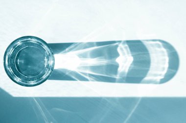 Temiz su ve keskin gölgeli bir bardak beyaz ahşap bir masanın üzerinde duruyor. Beyaz masa üzerinde su olan cam manzaralı.