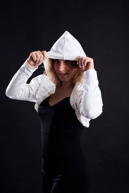 Genç sarışın kıvırcık kadın, siyah arka planda poz veriyor, gülümsüyor. Kadın portresinin dörtte üçü, beyaz bluz giyiyor, yüzü kapüşonlu. Işıklı karanlık anahtar fotoğrafçılığı.