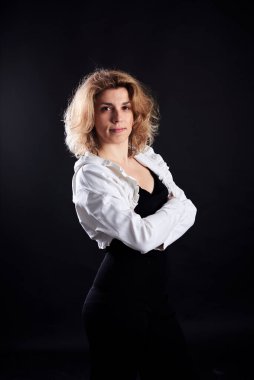 Genç sarışın kıvırcık kadın, siyah arka planda poz veriyor, gülümsüyor. Beyaz bluz giyen bir kadının dörtte üçü. Kendini sev konsepti. Işıklı karanlık anahtar fotoğrafçılığı.