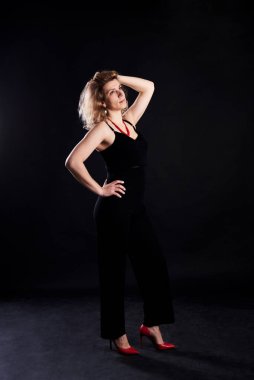 Siyah arka planda poz veren genç sarışın kıvırcık kadın. Işıklı fotoğraf stüdyosunda, siyah elbiseler ve kırmızı topuklu bir kadın portresi. Kendini sev.