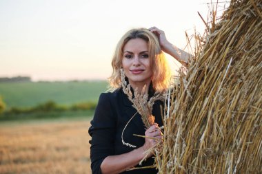 Siyah ceket ve kot pantolon giyen, yaz akşamları kırsal alanda buğday balyasına yaslanan genç sarışın kadın. Gün batımında doğal ortamda dörtte üç kadın portresi.