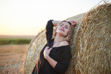 Siyah ceket ve kot pantolon giyen, yaz akşamları kırsal alanda buğday balyasına yaslanan genç sarışın kadın. Gün batımında doğal ortamda dörtte üç kadın portresi.
