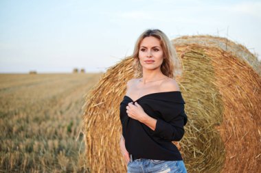 Siyah ceket ve kot pantolon giyen, yaz akşamları kırsal alanda buğday balyasının yanında duran genç sarışın kadın. Gün batımında doğal ortamda dörtte üç kadın portresi.