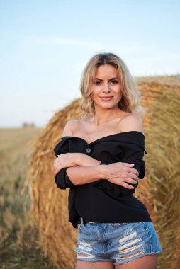 Siyah ceket ve kot pantolon giyen, yaz akşamları kırsal alanda buğday balyasının yanında duran genç sarışın kadın. Gün batımında doğal ortamda dörtte üç kadın portresi.