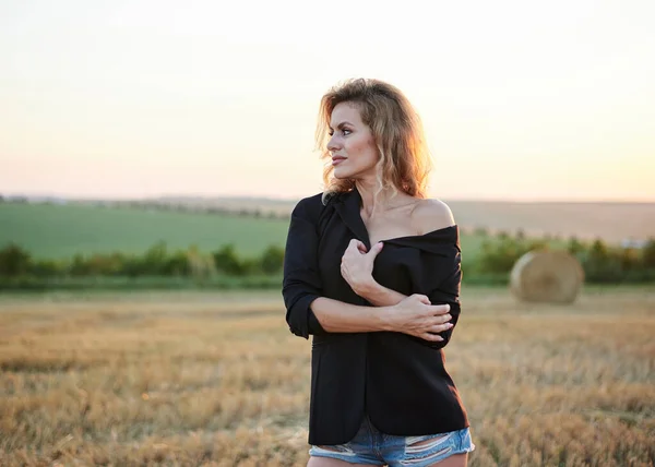 年轻的金发女人穿着黑色夹克和牛仔裤短裤 夏夜时分在麦田里摆姿势 日落时分 在自然的乡村风景中 创作时尚的四分之三女性肖像 — 图库照片