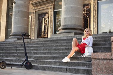 Şehir merkezinde sütunları olan eski tarihi binanın merdivenlerinde oturan genç sarışın kadın. Elektrikli scooterlı kadın portresi. Yaz tatili aktivitesi. Gezgin şehri keşfediyor.