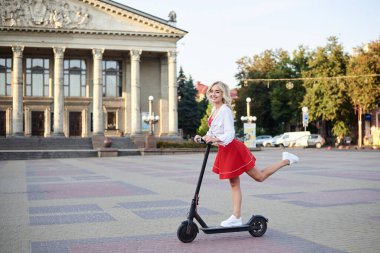 Şehir merkezinde, sütunları olan eski tarihi binanın önünde siyah elektrikli scooter kullanan genç sarışın bir kadın. Yaz tatili aktivitesi. Hava sıcakken dışarıda vakit geçirmek..