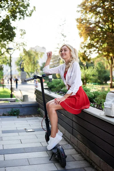 年轻的金发女子 坐在长椅上与电动滑板车在市中心 身穿红色裙子和白色上衣 摆出夏季休闲活动的姿势 乘摩托车游览市区的旅客 — 图库照片