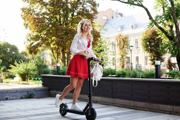 年轻的金发女人骑着电动车在市中心全长的女性肖像 身穿红色短裙和白色上衣 夏季休闲活动的娱乐活动 游客到城里游览 — 图库照片