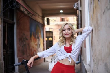Şehir merkezinde eski bina duvarının yanında elektrikli scooterlı genç sarışın kadın. Kadın, kırmızı ve beyaz giyinmiş, poz veriyor. Boş zaman etkinliği. Gezgin şehri keşfediyor..