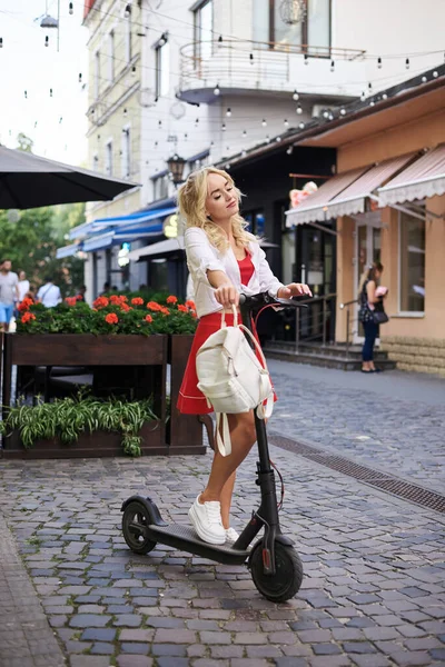 年轻的金发女人 站在市中心靠近花坛的地方 带着电动车 穿着红白相间衣服的女性 夏天的休闲活动 — 图库照片
