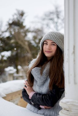 Genç esmer kız, gri kürklü ve örgü şapkalı, eski tarihi binanın balkonunda dikiliyor, sütunlara yaslanıyor. Dışarıdaki kadın portresi. Karlı şehir turnesinde turist..