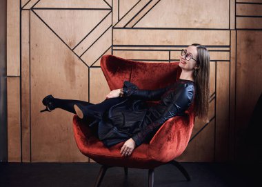 Süslü siyah deri elbise ve gözlük takan genç esmer kadın, ahşap duvarın önündeki kırmızı kadife Noel koltukta oturuyor, Mutlu Noeller ve mutlu yeni yıl kutlamaları. Kadın portresi