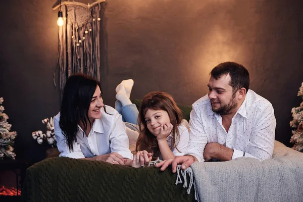 Mother. father and daughter, wearing white shirts, lying on bed in cozy dark room. Christmas and new year celebration with family. Winter holidays fun leisure time. Happy new year and Merry Christmas.