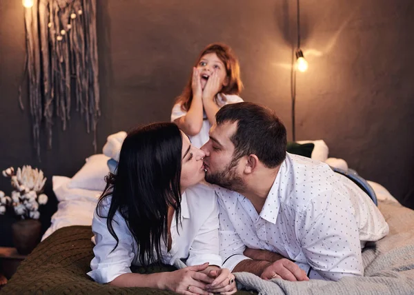 Mother. and father kissing and daughter making surprised face at the back of bed in cozy dark room. Christmas and new year celebration with family. Winter holidays fun leisure time.
