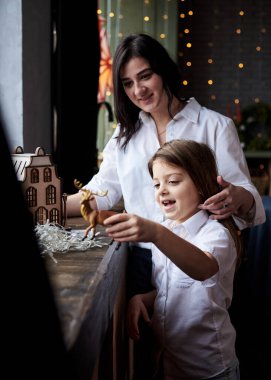 Anne ve kız, pencere kenarında dikilip, Noel süslemeleriyle karanlık odada tahta oyuncak ev ve geyiklerle oynuyorlar. Kış tatilleri evde. Karantina zamanında boş zaman aktivitesi.