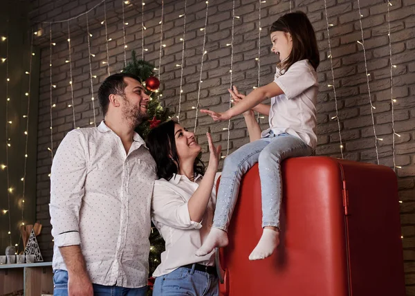 Small girl sitting on red fridge, giving hi five to her parents in dark kitchen with christmas lights. Mother, father and daughter spending winter holidays at home during quarantine.Family activities