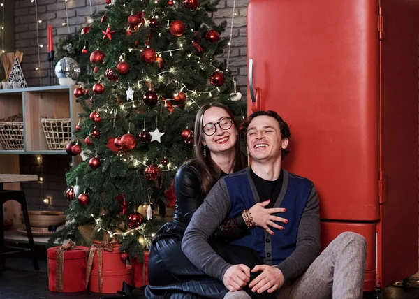 Young married couple sitting on kitchen floor near red fridge and Christmas tree Man and woman celebrating winter holidays at home during quarantine. Happy new year. Holiday weight gaining.