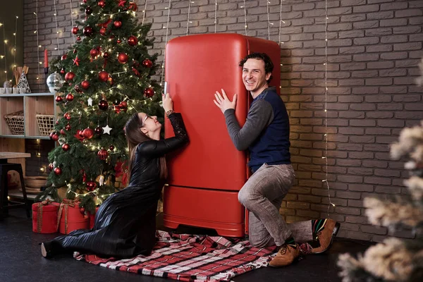 Young married couple kneeling down on kitchen floor near red fridge, begging for food. Man and woman celebrating winter holidays at home during quarantine.Happy new year. Holiday weight gaining.