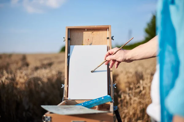Close-up picture of hands of male artist, drawing on white canvas on wheat field in summer. Painting workshop in rural countryside. Artistic education concept. Outdoors leisure activities.