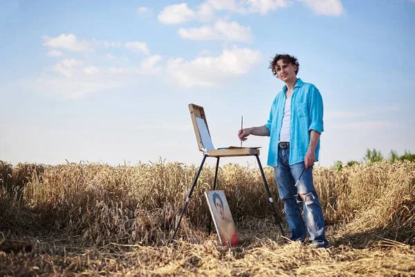 Full-length portrait of young male artist wearing blue shirt, drawing on canvas on easel on wheat field. Painting workshop in rural countryside. Artistic education concept. Outdoors leisure activities