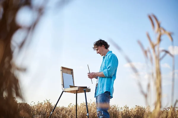 Young male artist wearing light blue shirt, drawing on canvas on sketchbook easel on wheat field. Painting workshop in rural countryside. Artistic education concept. Outdoors leisure activities.