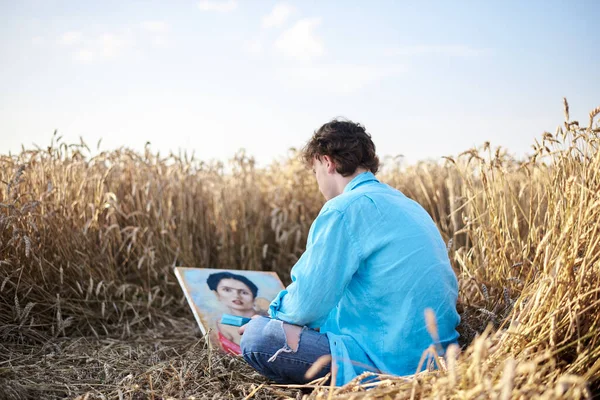 Young male artist, wearing blue shirt and torn jeans, sitting on wheat field, drawing on canvas. Painting workshop in rural countryside. Artistic education concept. Outdoors leisure activities.