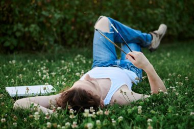 Genç erkek sanatçı, yırtık kot pantolon ve beyaz tişört giyiyor, gün batımında yeşil çimlerin üzerinde yatıyor, fırça tutuyor, yanında boş bir tuval ile ne çizeceğini düşünüyor. Sanatsal ilham kavramı.