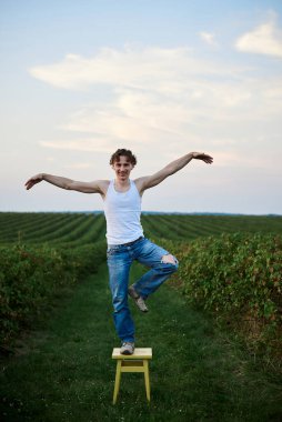 Genç yakışıklı bir adam, yırtık kot pantolon ve beyaz tişört giyiyor, yeşil alanda sarı taburede duruyor, eğleniyor. Sanatsal eğitim konsepti. Dışarıda yaratıcı bir portre.