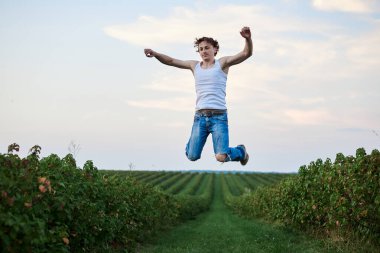 Genç yakışıklı adam, yırtık kot pantolon ve beyaz tişört giyiyor, yeşil alanda havaya zıplıyor, gülümsüyor, gülüyor, eğleniyor. Sanatsal eğitim konsepti. Dışarıda yaratıcı bir portre var. Özgürlük ilham kaynağı.