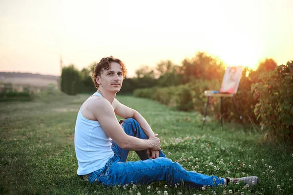 Young male artist, wearing torn jeans and white t-shirt, sitting on ground on green field during sunset, with painting on background. Painting workshop. Artistic inspiration concept.