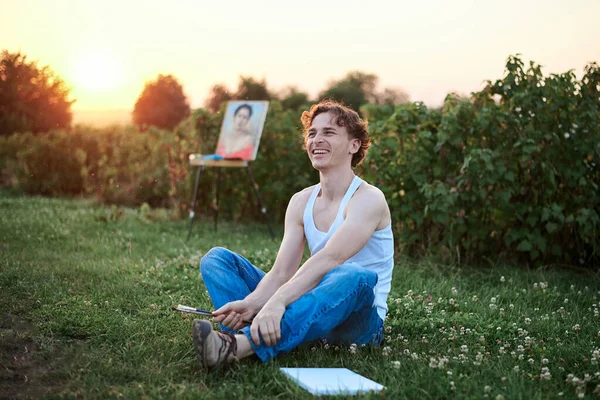 Young male artist, wearing torn jeans and white t-shirt, sitting on ground on green field during sunset, with painting on background, smiling. Painting workshop outside. Artistic inspiration concept.