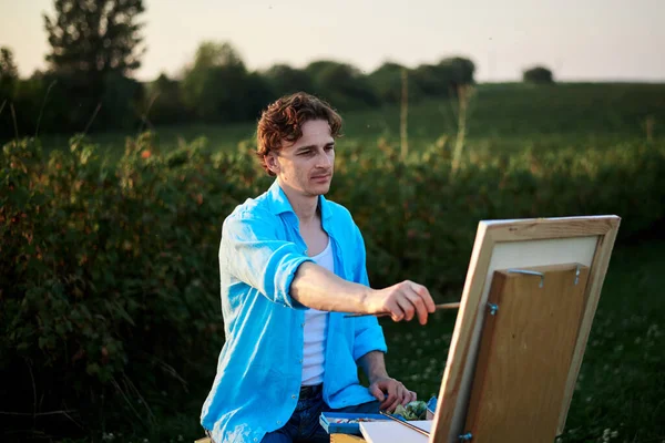 Young male artist, sitting by easel, drawing portrait on canvas on green field in summer. Painting workshop in rural countryside. Artistic education modern concept. Outdoors leisure activities.