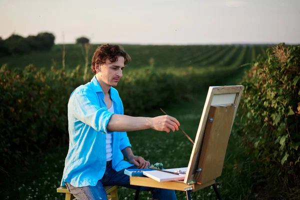 Young male artist, sitting by easel, drawing portrait on canvas on green field in summer. Painting workshop in rural countryside. Artistic education modern concept. Outdoors leisure activities.