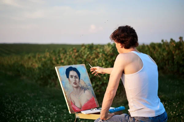 Young male artist, sitting by easel, drawing portrait on canvas on green field in summer. Painting workshop in rural countryside. Artistic education modern concept. Outdoors leisure activities.