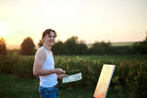 Young male artist, standing by easel, drawing portrait on canvas on green field in summer during sunset. Painting workshop in countryside. Artistic education modern concept.Outdoors leisure activity.