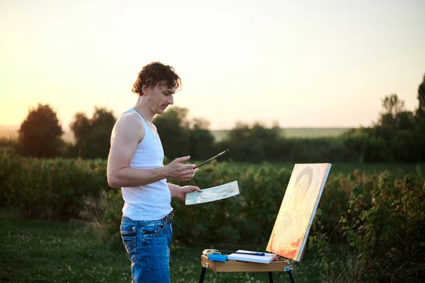 Young male artist, standing by easel, drawing portrait on canvas on green field in summer during sunset. Painting workshop in countryside. Artistic education modern concept.Outdoors leisure activity.