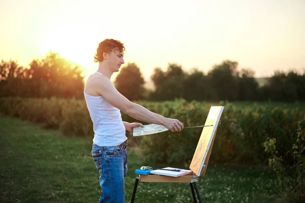 Young male artist, standing by easel, drawing portrait on canvas on green field in summer during sunset. Painting workshop in countryside. Artistic education modern concept.Outdoors leisure activity.