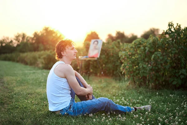 Young male artist, wearing torn jeans and white t-shirt, sitting on ground on green field during sunset, with painting on background. Painting workshop. Artistic inspiration concept.