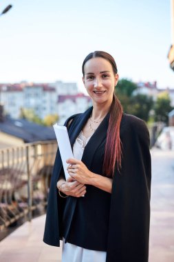 Siyah ceketli, beyaz elbiseli, elinde kağıtlarla balkonda duran esmer bir iş kadını. Yazın kadın portresi. Terasta güzel şehir manzaralı bir model..