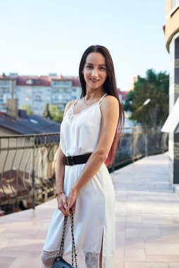 Genç esmer kadın, ipek beyaz elbise giyiyor, siyah çanta taşıyor, sabah otel balkonunda duruyor. Yazın kadın portresi. Terasta poz veren güzel şehir manzaralı manken..