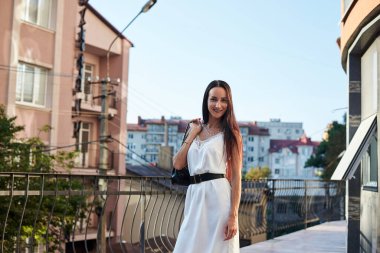 Genç esmer kadın, ipek beyaz elbise giyiyor, siyah çanta taşıyor, sabah otel balkonunda duruyor. Yazın kadın portresi. Terasta poz veren güzel şehir manzaralı manken..