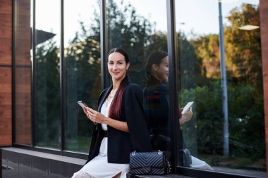 Kırmızı at kuyruklu esmer kız, şık siyah ceket ve beyaz ipek elbise giyiyor, modern cam binanın yanında oturuyor, telefonu tutuyor. Yemek arası vermiş güzel bir iş kadını. Kadın portresi