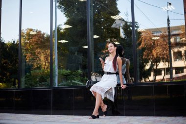 Kırmızı at kuyruklu esmer kız, beyaz ipek elbise giyiyor, modern cam binanın yanında oturuyor, telefonu tutuyor. Yemek arası vermiş güzel bir iş kadını. Kadın şehir portresi