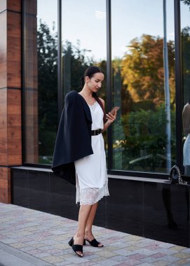 Kırmızı at kuyruklu esmer kız, şık siyah ceket ve beyaz ipek elbise giyiyor, modern cam binanın yanında duruyor, telefonu tutuyor. Yemek arası vermiş güzel bir iş kadını. Kadın portresi