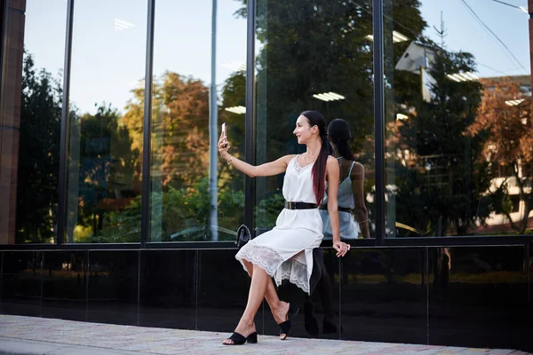 Young brunette girl with red pony tail, wearing white silk dress, sitting near modern glass building, taking selfie with phone. Pretty business woman on lunch break. Female city portrait