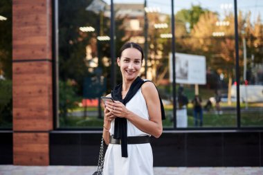 Kırmızı at kuyruklu esmer kız, şık beyaz ipek elbise giyiyor, cam binanın önünde telefon tutuyor. Yemek arası vermiş güzel bir iş kadını. Romantik kadın şehir portresi.