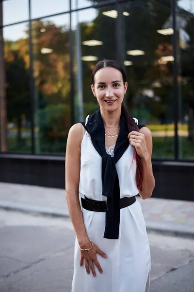 Young brunette girl with red pony tail, wearing stylish white silk dress and black blouse, standing in front of glass building. Pretty business woman on lunch break. Romantic female urban portrait.