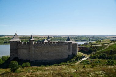 Nehirle çevrili antik kalenin manzarası. Mavi güneşli gökyüzü ve parlak sarı bitkilerle güzel bir doğa manzarası. Yaz tatili. Tarihi gezi turu
