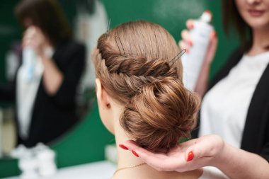 Gelin düğüne hazırlanıyor. Profesyonel kuaför kadın müşteriye kuaförlük yapıyor, büyük aynanın önüne saç spreyi sıkıyor. Güzellik stüdyosunda çalışma süreci. Gala saç modelinin yakın plan fotoğrafı..